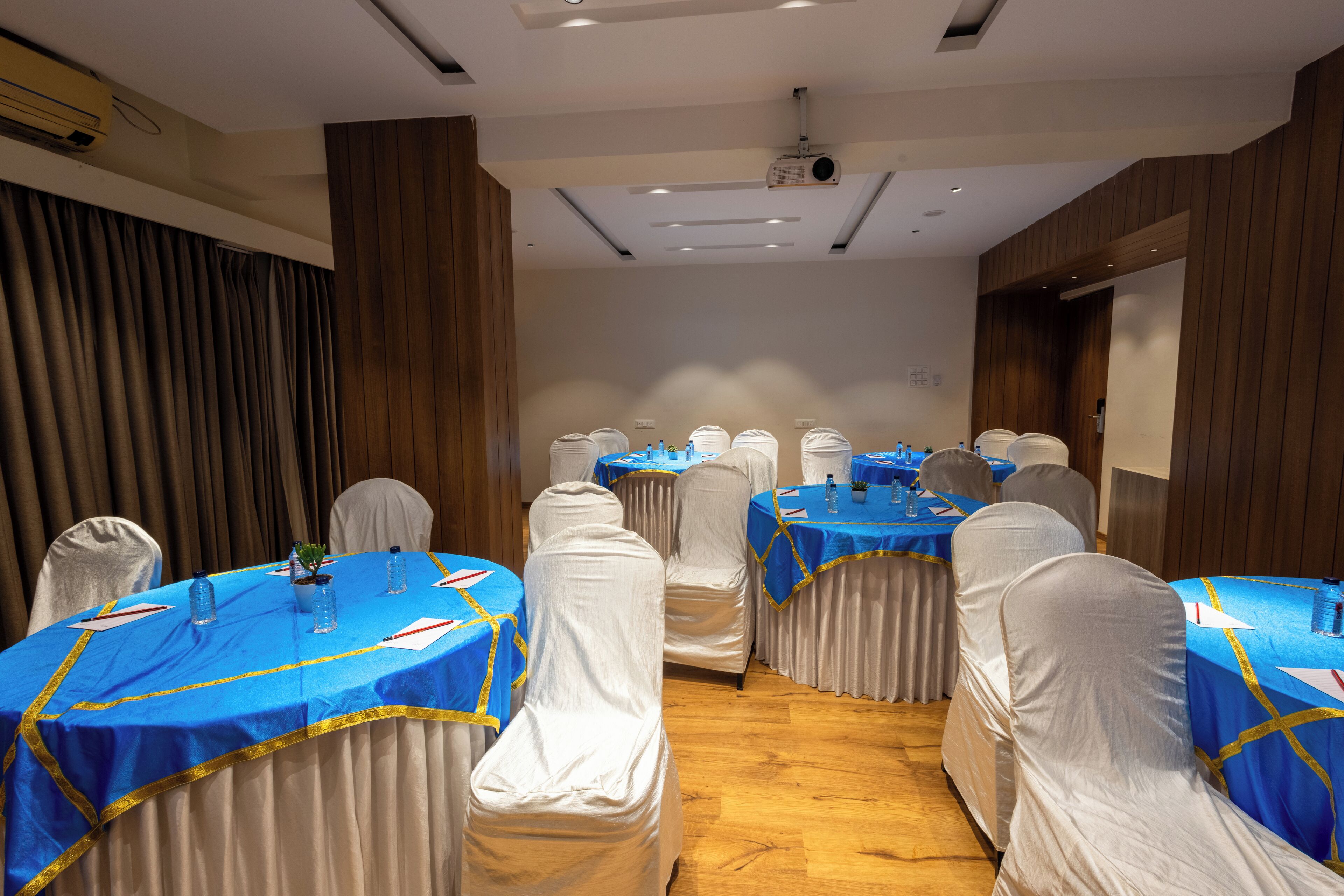 Una sala conferenze con tavoli rotondi ricoperti da tovaglie blu, sedie bianche e uno schermo per proiettore sulla parete.