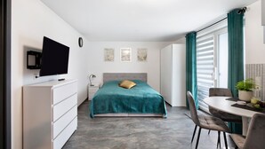A bedroom with a bed, a TV, a dresser, a bedside table, a chair, and a window with curtains.