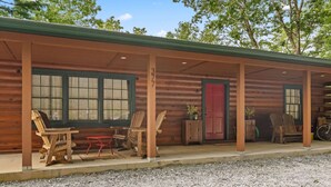 A wooden cabin with a covered porch, two wooden chairs, a small table, and a red door.