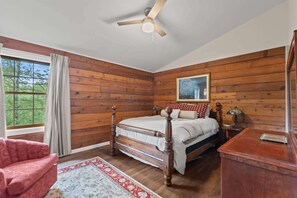 A bedroom with a wooden paneled wall, a bed with a headboard, a nightstand, a lamp, and a window with curtains. - Little Birds Nest (Highlands)