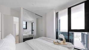 A modern bedroom with a large bed, a bedside table with a vase and cups, and a view of the city through large windows. - Spacious 2BR City Apt, Panoramic Views & Parking (Southbank)