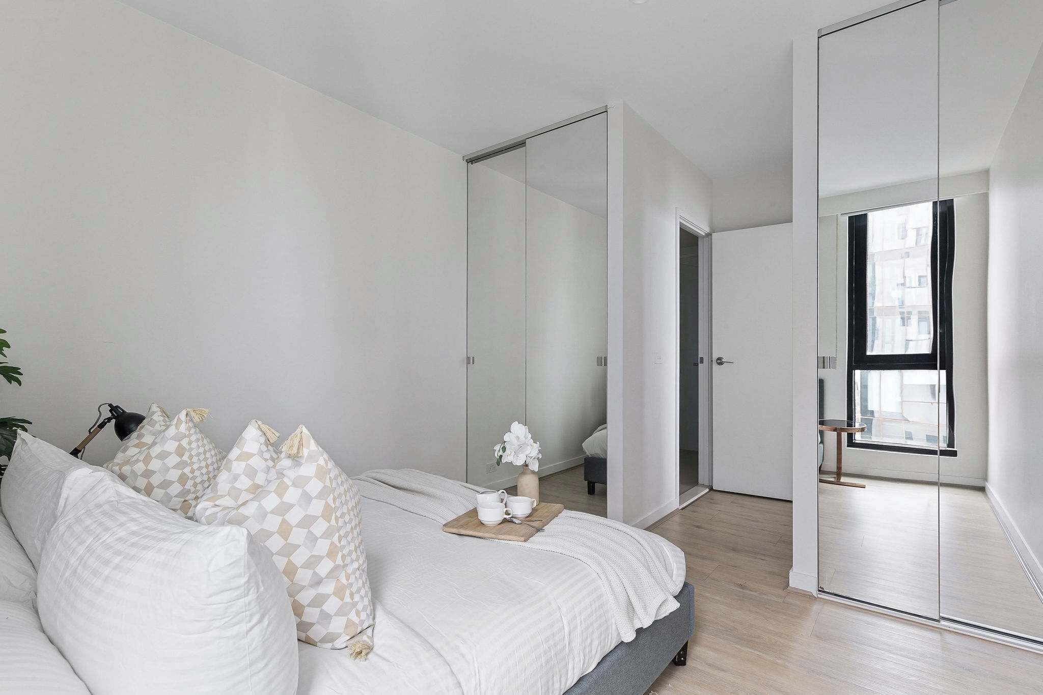 A modern bedroom with a bed, white bedding, a bedside table with a vase of flowers, and a large mirror.
