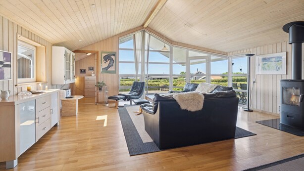 A spacious living area with a wooden ceiling, a black sofa, a fireplace, and large windows offering a view of the outdoors.