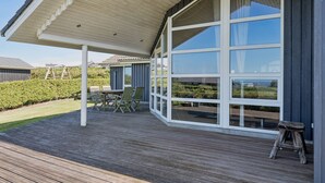 A covered outdoor deck with a table and chairs, large windows, and a wooden stool.