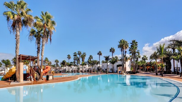 A swimming pool with palm trees, a water slide, and a building with a red roof. - Oasis Duna 'Casa Martineli' with Shared Pool, Balcony, and Wi-Fi (Corralejo)
