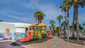 A playground with slides and climbing structures, palm trees, and a mural on a building. - Oasis Duna 'Casa Martineli' with Shared Pool, Balcony, and Wi-Fi (Corralejo)