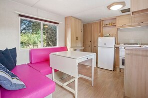 A compact living space with a kitchenette, a dining table, a sofa, and a window with a view of trees. - Mobile Home 'Kenya' with Shared Pool, Wi-Fi and Air Conditioning (Le Val)