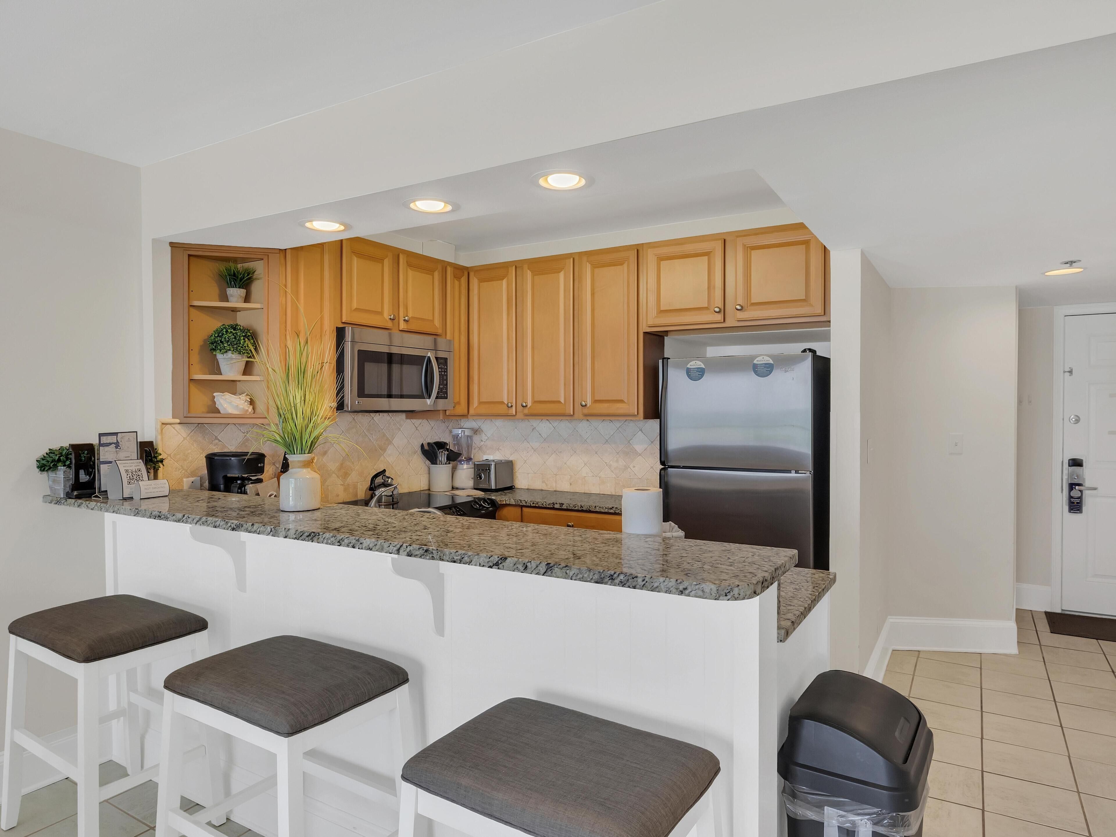 A modern kitchen with wooden cabinets, a stainless steel refrigerator, a microwave, and a granite countertop. There are three bar stools with cushioned seats.