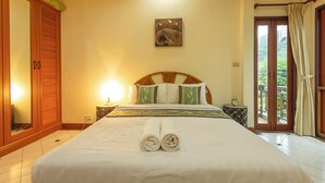 A neatly made bed with rolled towels, a wooden headboard, a bedside table with a lamp, and a view of the outdoors through a sliding door. - Thai-Style Villa with Charm V114 (Kamala)