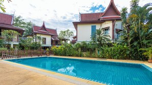 A swimming pool with a tiled edge, surrounded by tropical plants and traditional buildings with red roofs. - Thai-Style Villa with Charm V114 (Kamala)