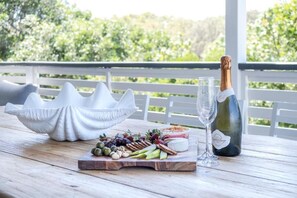 Et træbord med et ostebræt, druer, nødder og en champagneflaske på en balkon med gelænder og grøn vegetation i baggrunden.