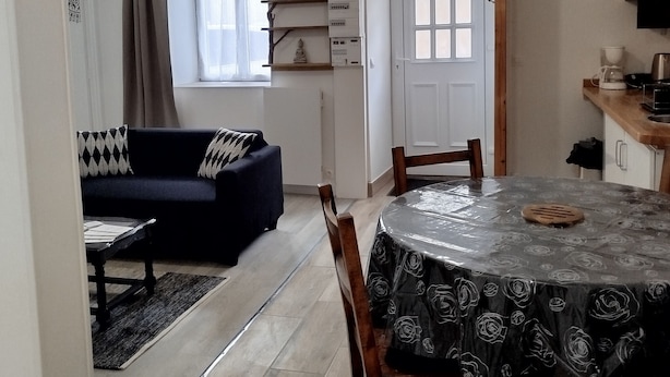 A dining area with a round table covered by a rose-patterned tablecloth, wooden chairs, and a coffee machine on the counter. - Renovated apartment in quiet wine-growing village. 15min from Auxerre. (Irancy)