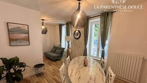 A dining area with a marble table and white chairs, a hanging light fixture, and a view of the outdoors through a window. - La Parenthèse Douce (Saint-Brieuc)
