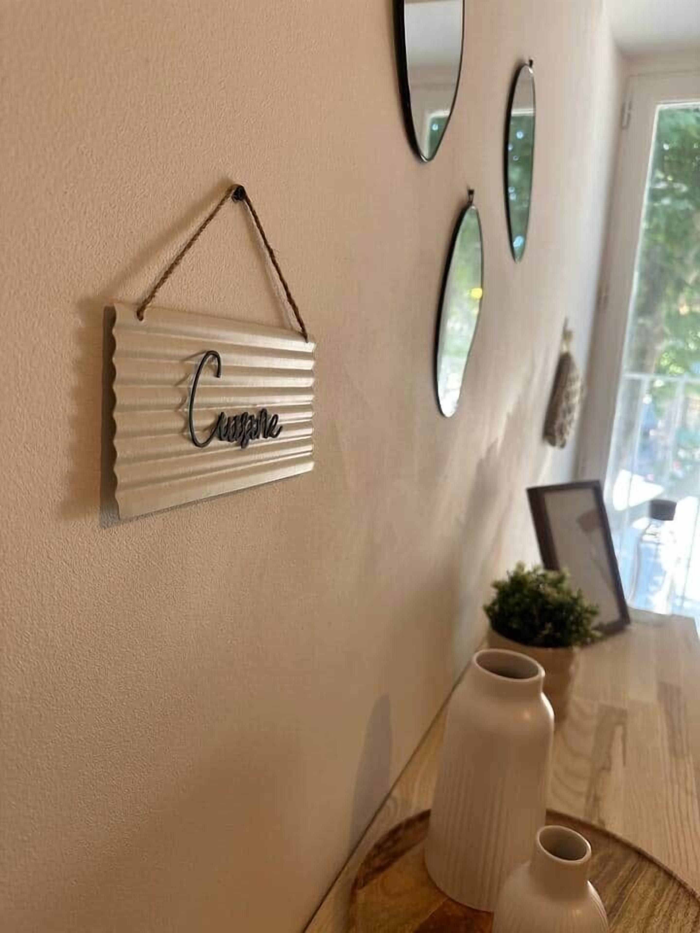 Una pared con un letrero metálico colgante que dice "Cousine", tres espejos ovalados y una pequeña planta en maceta.