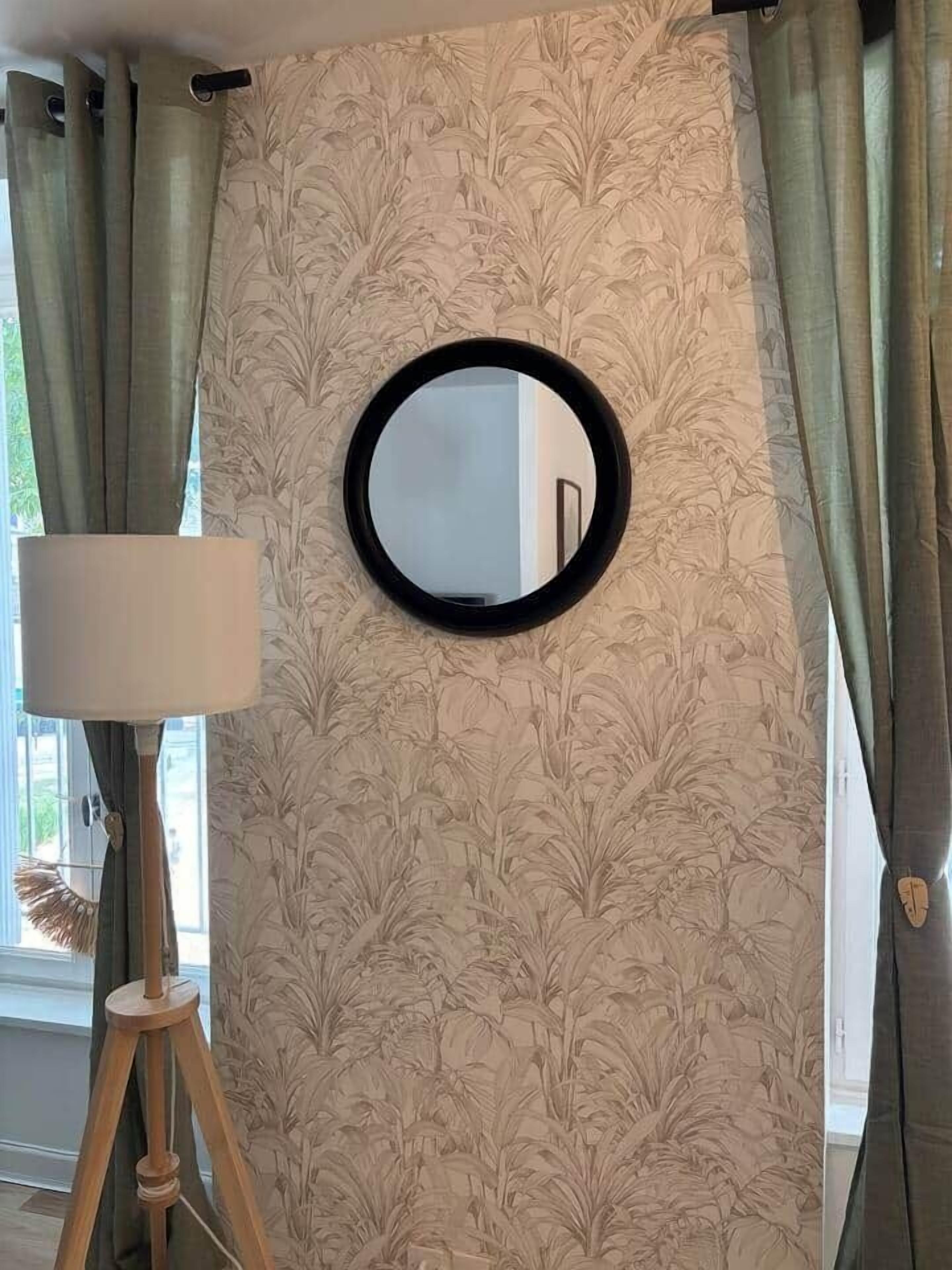 Une pièce avec un grand miroir rond accroché au mur, une lampe posée sur un trépied et des rideaux à motifs floraux.
