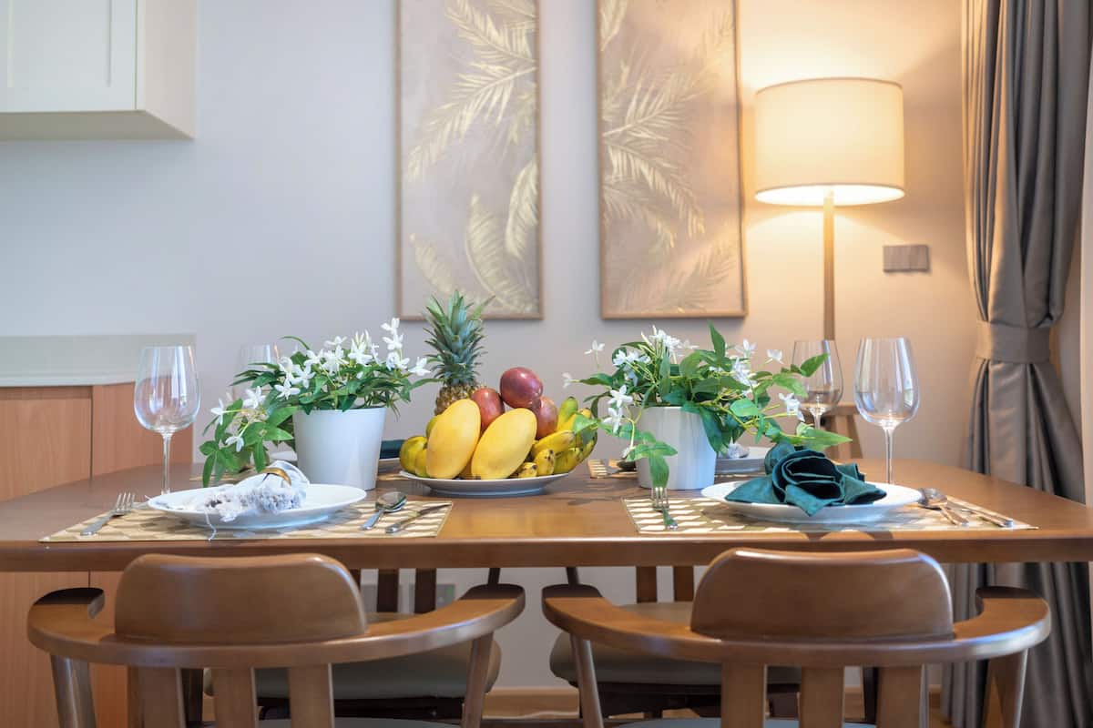 Une table à manger avec un panier de fruits, deux plantes en pot et une lampe.