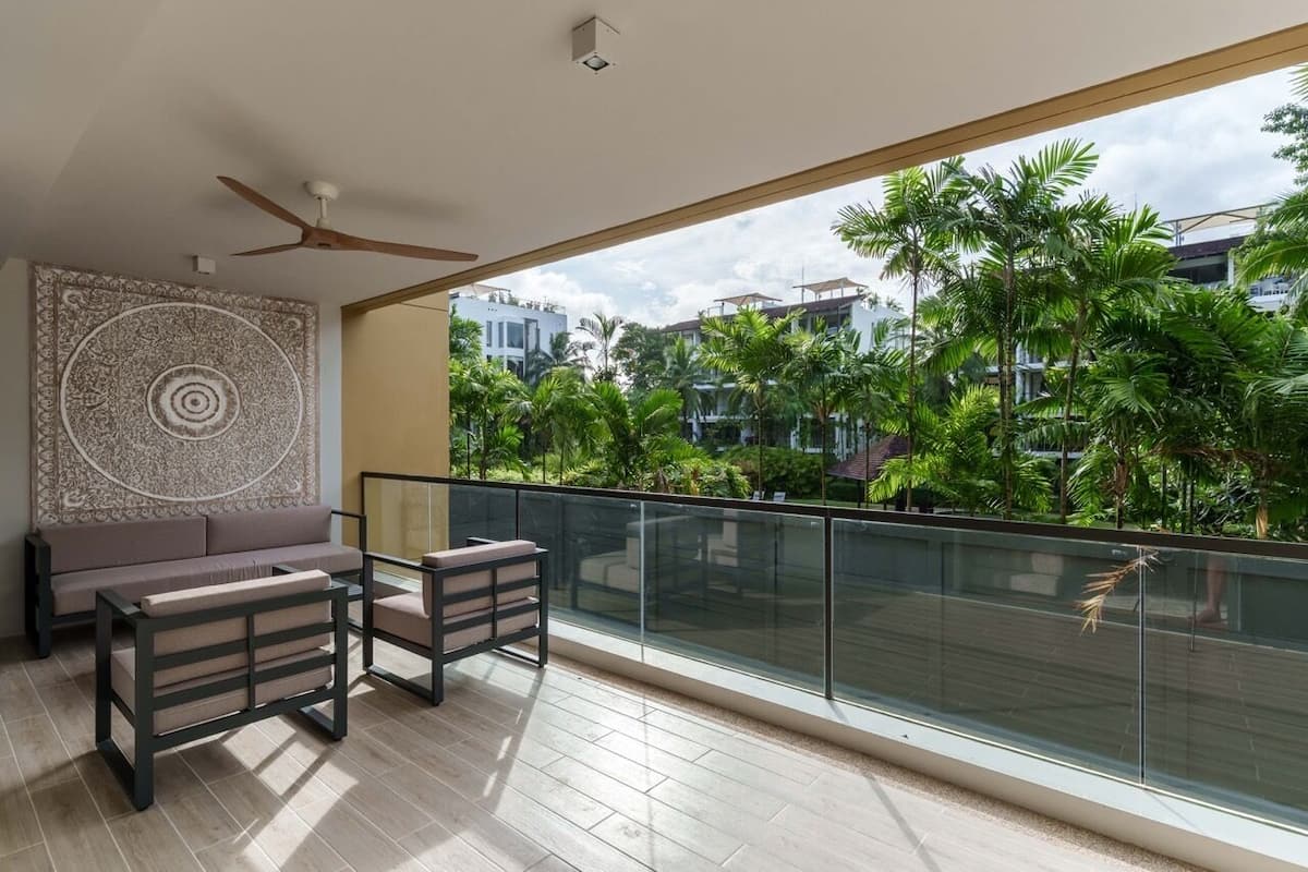 Un balcon avec un canapé et des fauteuils, un ventilateur de plafond et une vue sur les palmiers et les immeubles.