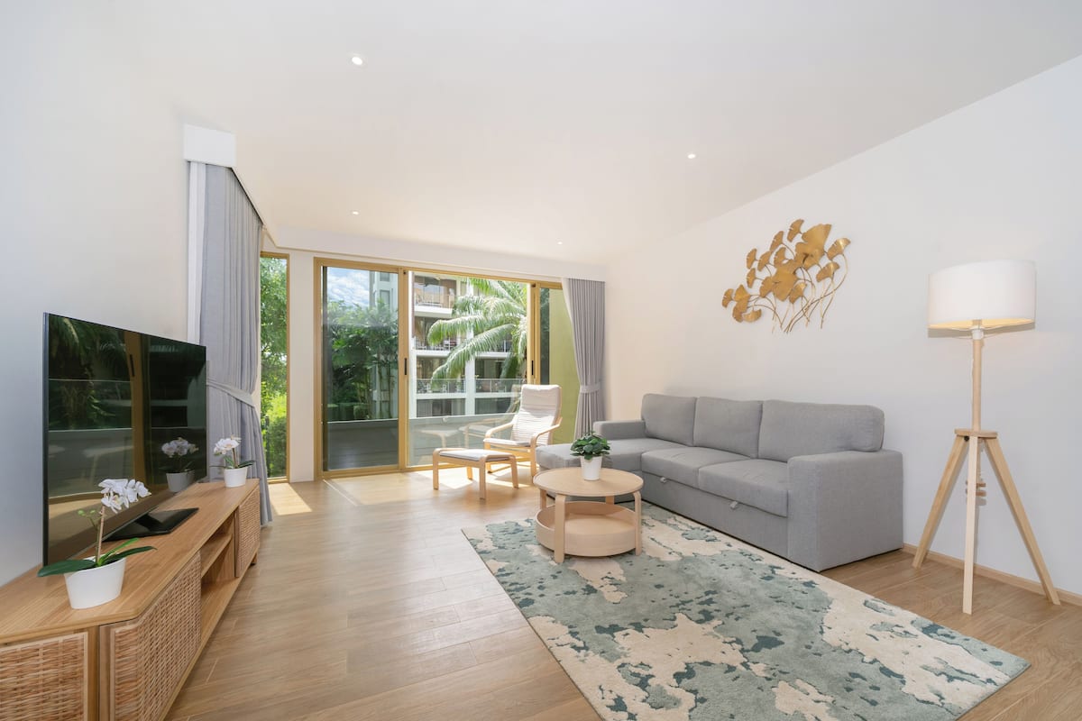 Un salon moderne avec un canapé gris, une table basse en bois, un téléviseur et une grande fenêtre donnant sur un paysage verdoyant.