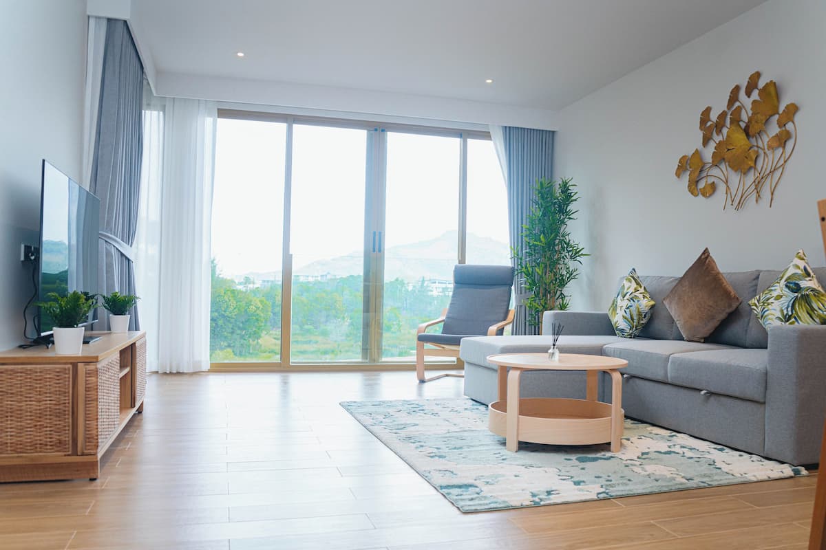 Un salon moderne comprenant un canapé, un fauteuil, une table basse, un téléviseur et une grande fenêtre donnant sur un paysage verdoyant.