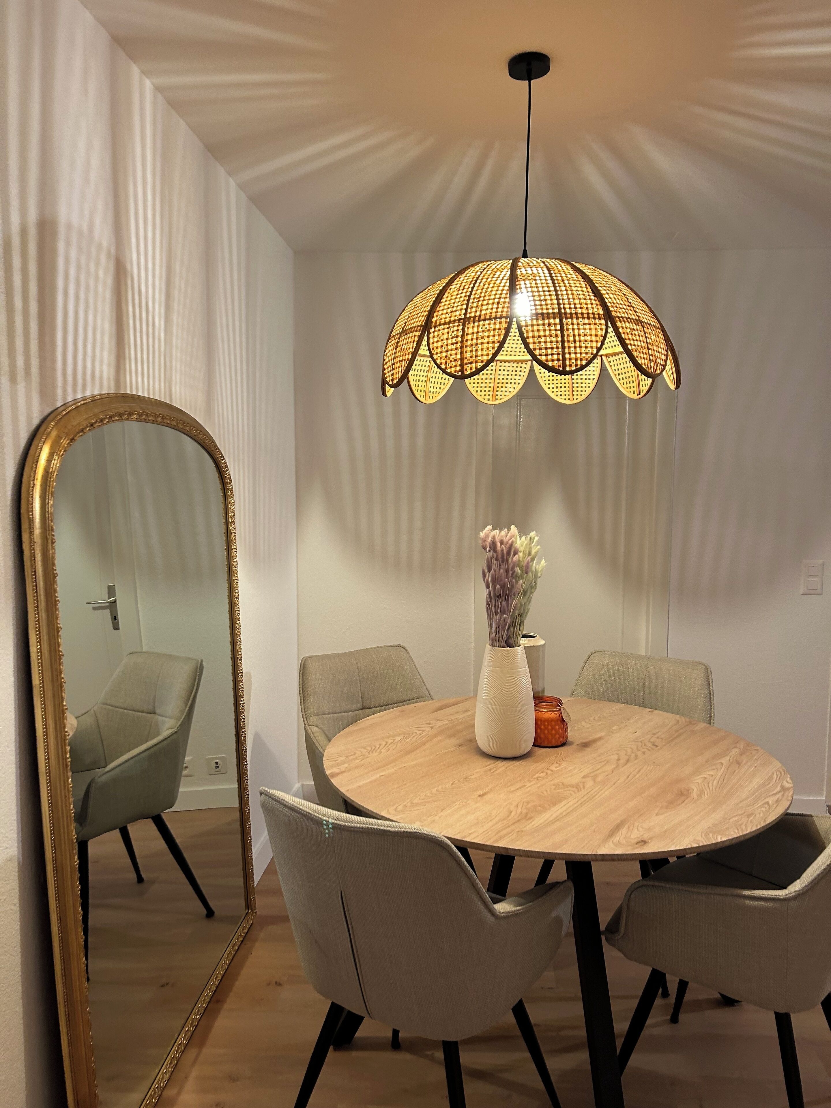 Un comedor con una mesa redonda de madera, cuatro sillas grises, un espejo grande con marco dorado y una lámpara colgante con pantalla de mimbre.