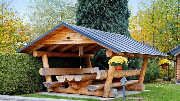 A rustic wooden structure with a sloped roof, logs stacked in front, and potted yellow flowers. - Teichmühle – Lakefront Apartment in Vogtland for Family & Pet-Friendly Holidays (Bad Brambach)