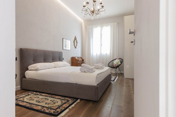 Une chambre à coucher moderne avec un grand lit, un lustre et une vue sur l’extérieur à travers des rideaux transparents.