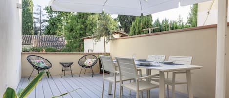 Une terrasse extérieure avec une table et des chaises blanches, un grand parasol et une vue sur un quartier résidentiel avec des arbres et des maisons.