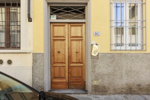 Une porte à deux battants en bois, surmontée d’une grille métallique, le numéro « 93 » peint sur le mur à droite, et une voiture garée devant.