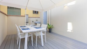 Un coin repas avec une table blanche dressée pour quatre personnes, un parasol blanc et une plante en pot.