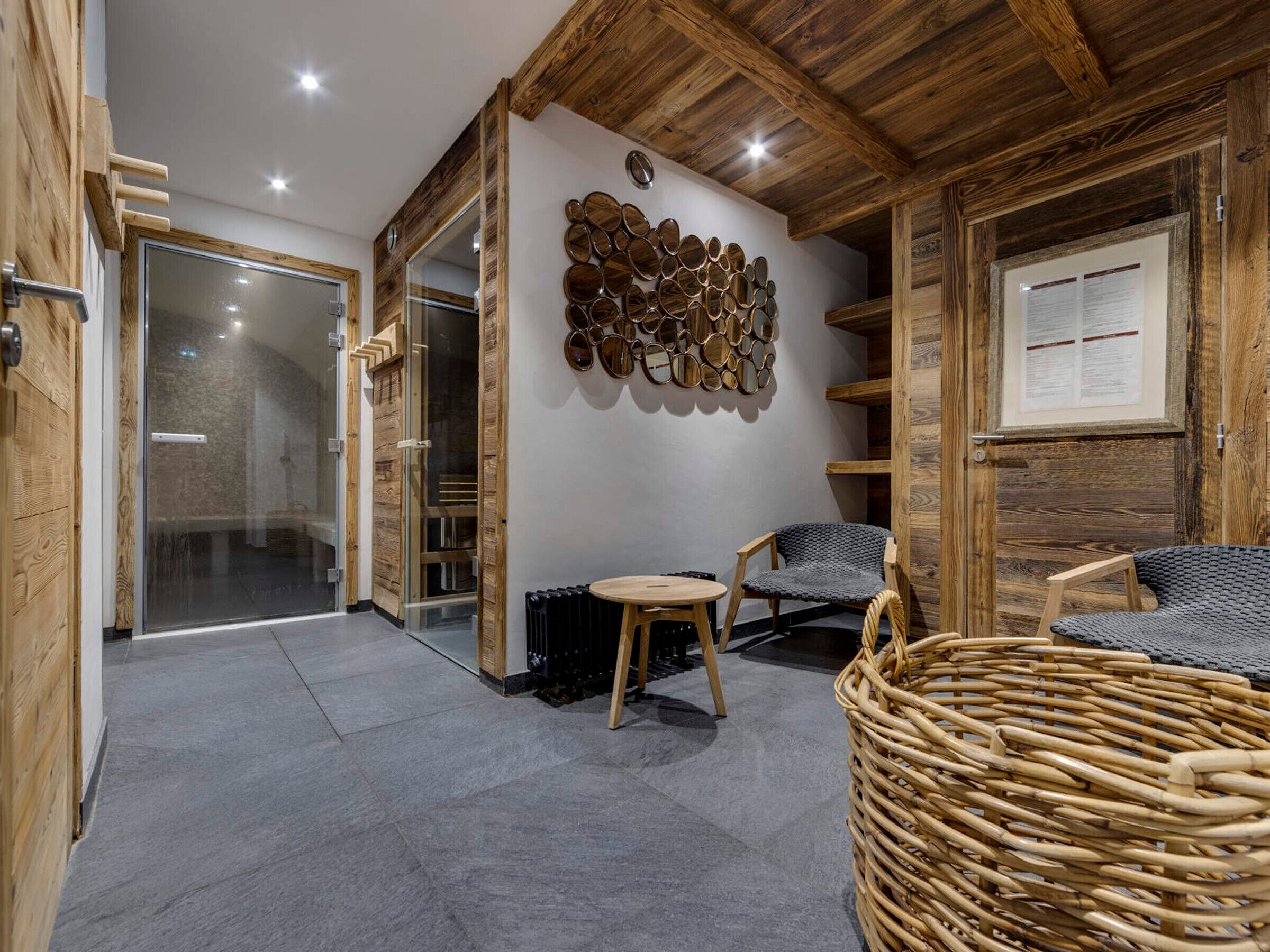 A room with a wooden ceiling, a wall-mounted mirror art piece, a wicker chair, and a small table.