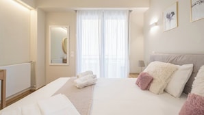 Quarto de hotel com cama grande, mesa de cabeceira, espelho e janela com cortinas leves.