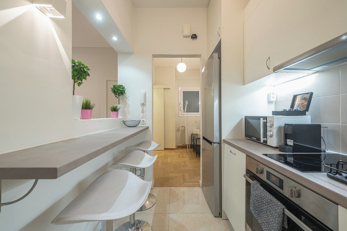 A modern kitchen with a built-in oven, microwave, and a digital clock. There are three white bar stools and potted plants on the counter.