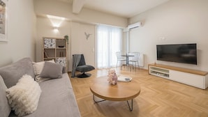 Uma sala de estar moderna com um sofá cinza, uma mesa de centro redonda, uma televisão de tela plana e um espaço de jantar com mesa e cadeiras.