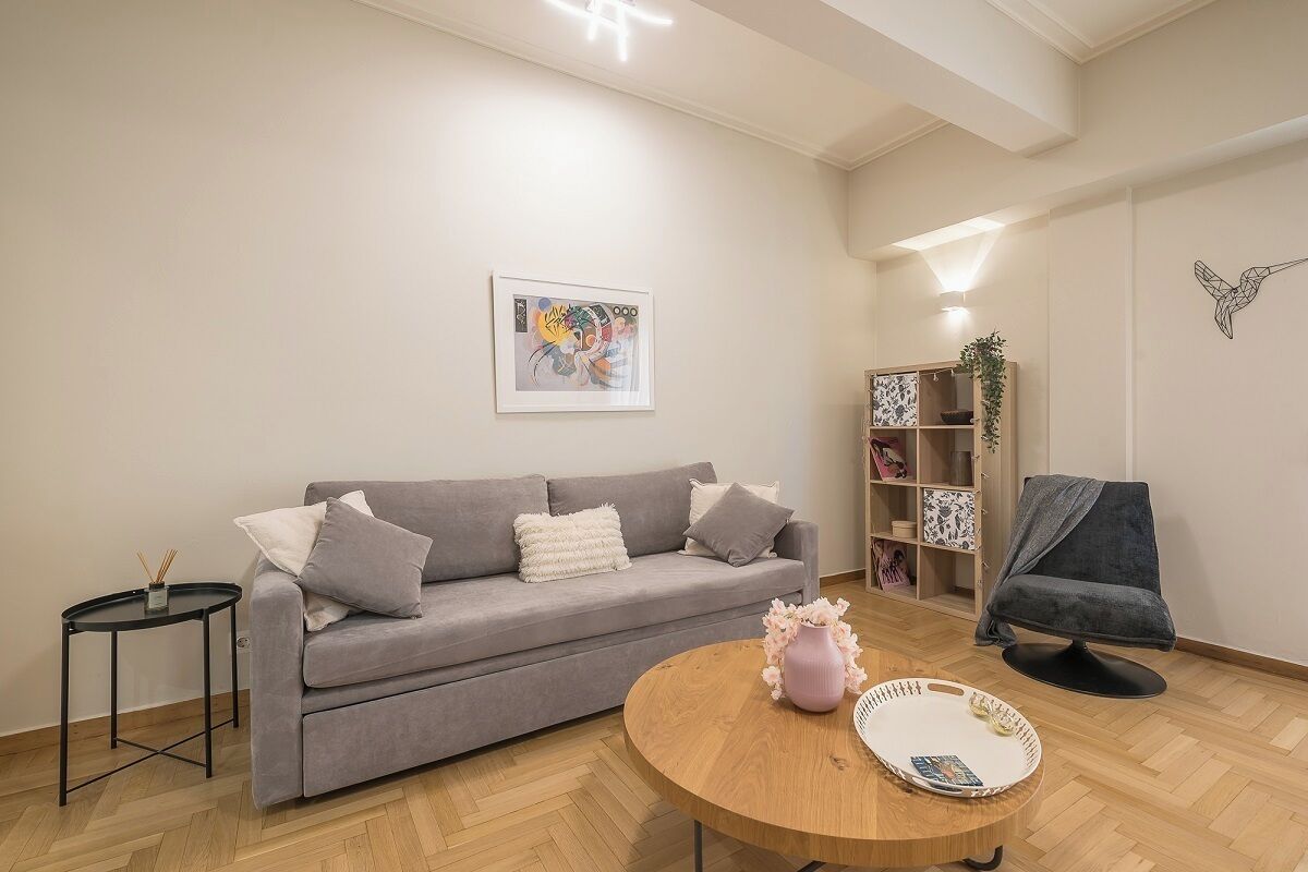 A living room with a grey sofa, a round wooden coffee table, a small black side table, a bookshelf, and a chair with a blanket.