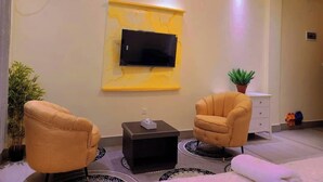 A modern living room with a flat-screen TV mounted on a yellow accent wall, two armchairs, a small side table, and a potted plant. - Hotel Santa Bella (Douala)
