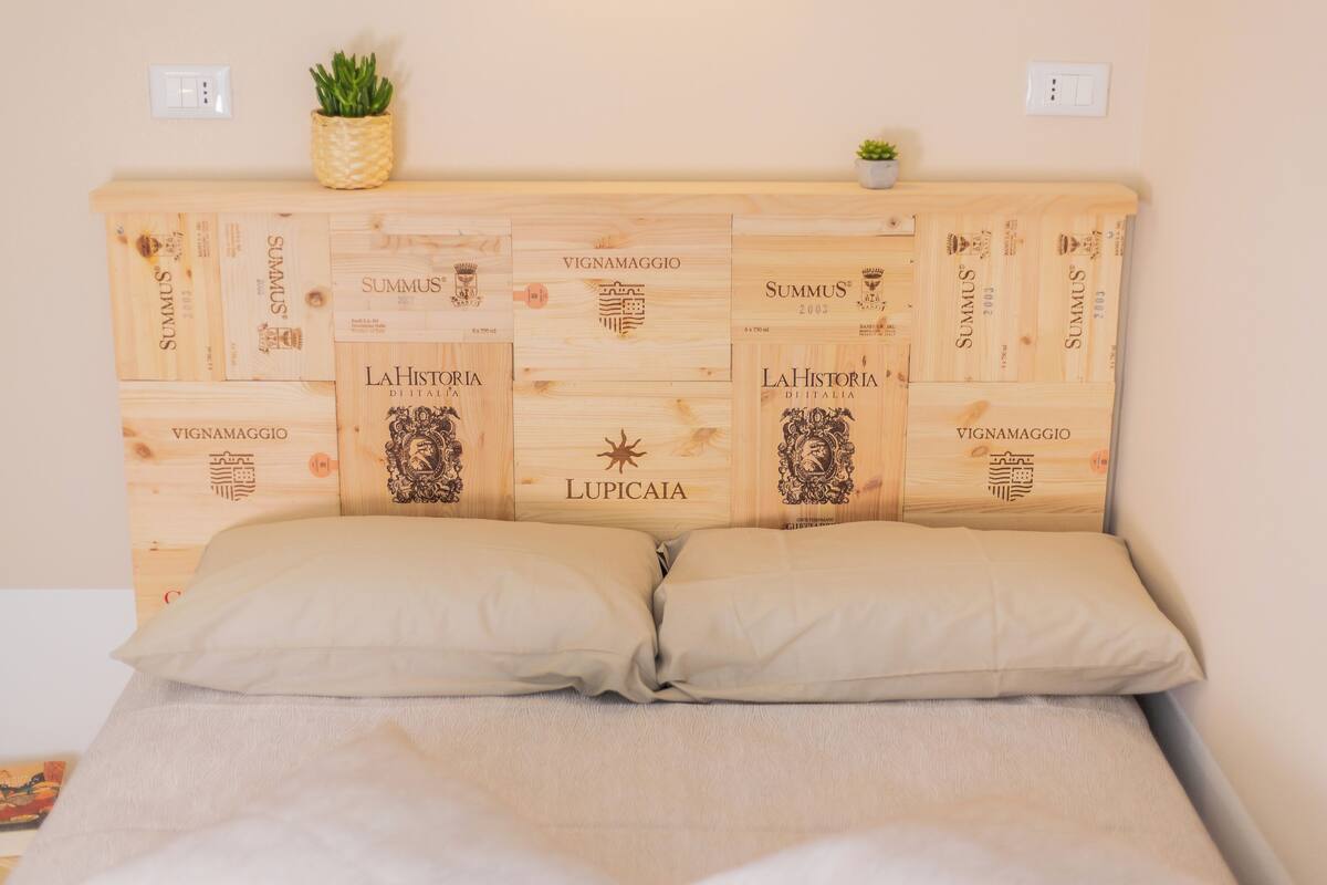 Una cama con cabecera de madera con inscripciones y logotipos, dos almohadas, una planta pequeña y un enchufe en la pared.