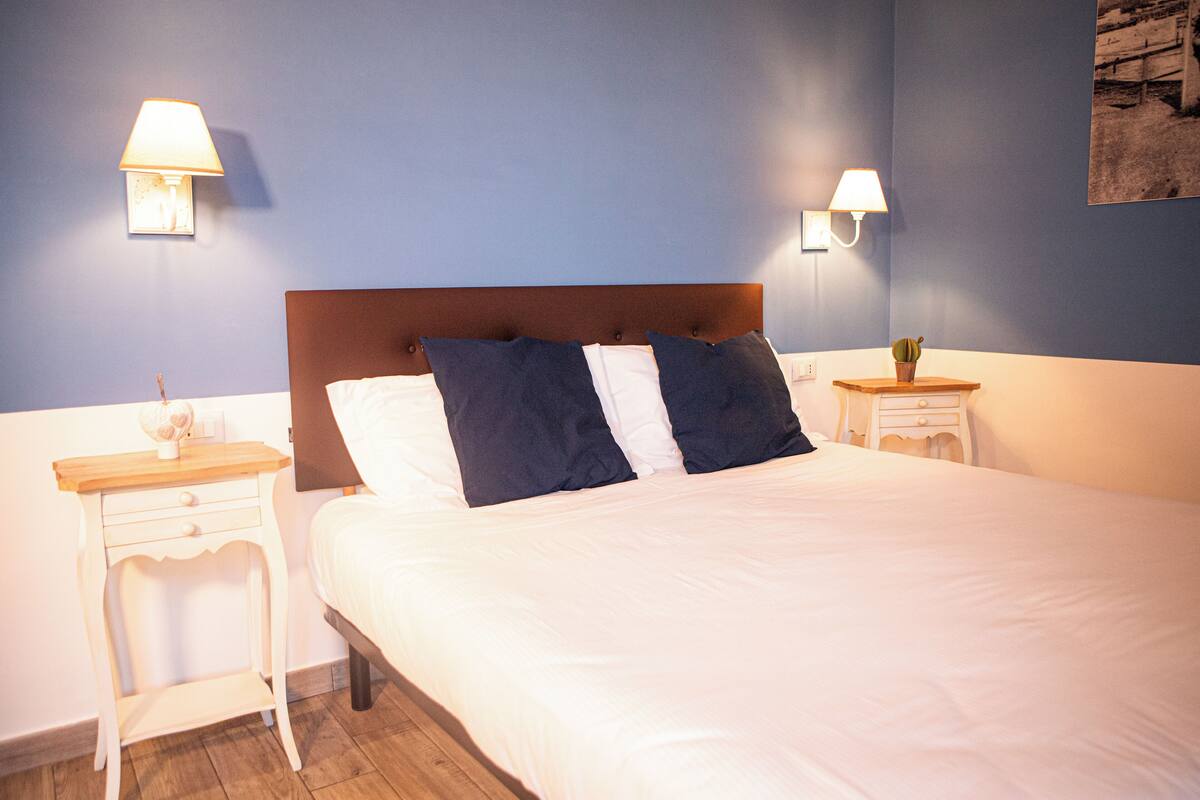 Una cama bien tendida con almohadas blancas y azul oscuro, dos mesitas de noche con lámparas y una pequeña planta en una de ellas.