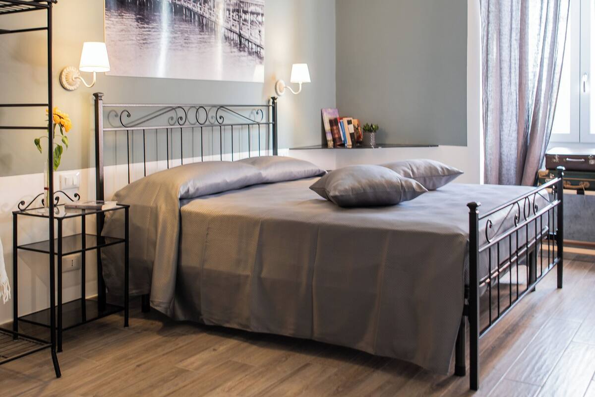 Un dormitorio con una cama, una mesita de noche, una lámpara, una ventana con cortinas y un cuadro colgado en la pared.