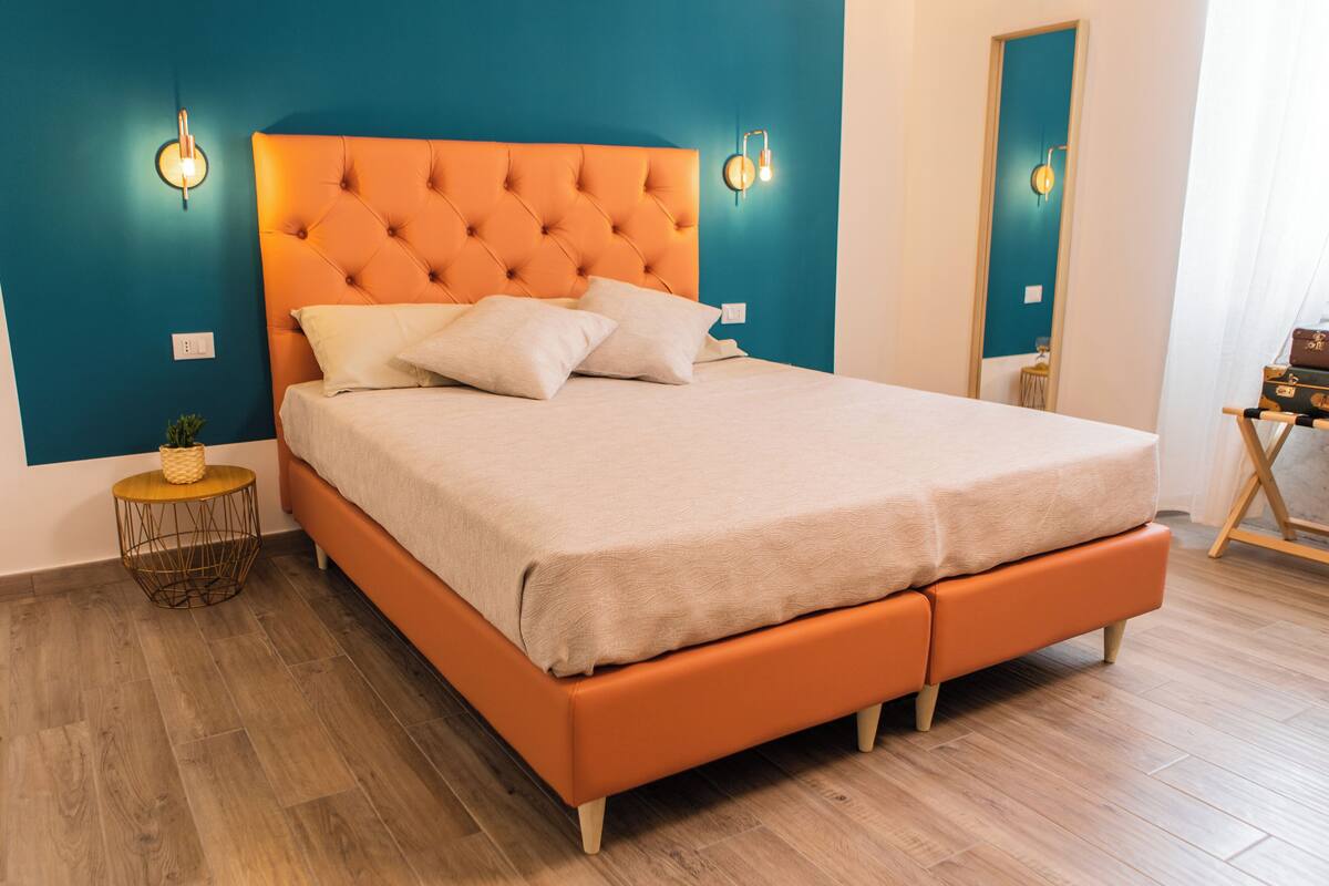 Una cama con cabecera naranja y ropa de cama blanca, una mesita redonda pequeña con una cesta, y suelo de madera.