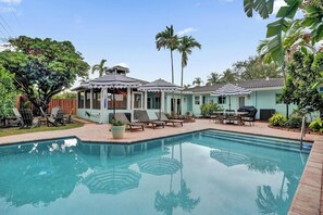 A swimming pool with lounge chairs and umbrellas, surrounded by palm trees and a covered patio area.