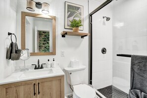 Un baño moderno con un lavamanos blanco, un espejo y un inodoro. Cuenta con una zona de ducha con puerta de vidrio y un perchero con toalla.
