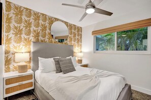Une chambre avec un lit, des tables de chevet, un ventilateur de plafond, une fenêtre donnant sur des palmiers et un papier peint à motifs d’ananas.