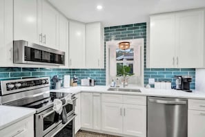 A modern kitchen with white cabinets, a stainless steel microwave, a built-in oven, a dishwasher, and a window with a view of greenery. - Poolside Palms Villa 🌴 Resort-Style Pool | Near Beach 🏖️ (Fort Lauderdale)