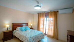 Een slaapkamer met een bed, nachtkastjes, een plafondventilator, een airconditioner en een raam met gordijnen.