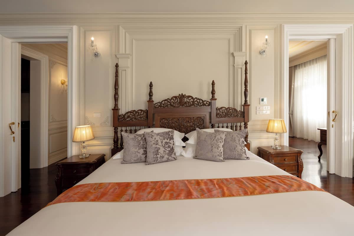A large bed with ornate headboard, two bedside tables with lamps, and a view of a hallway.