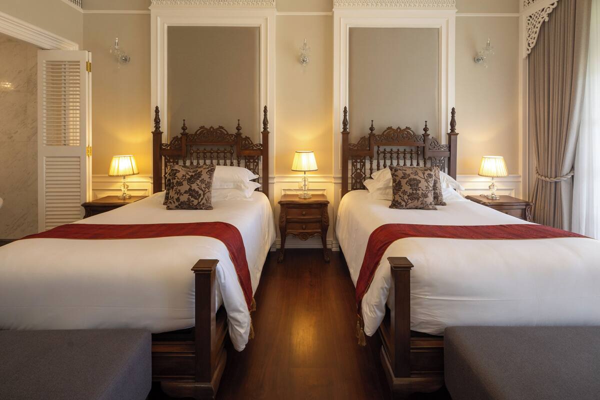 A hotel room with two beds, wooden headboards, bedside tables with lamps, a mirror, and a window with curtains.