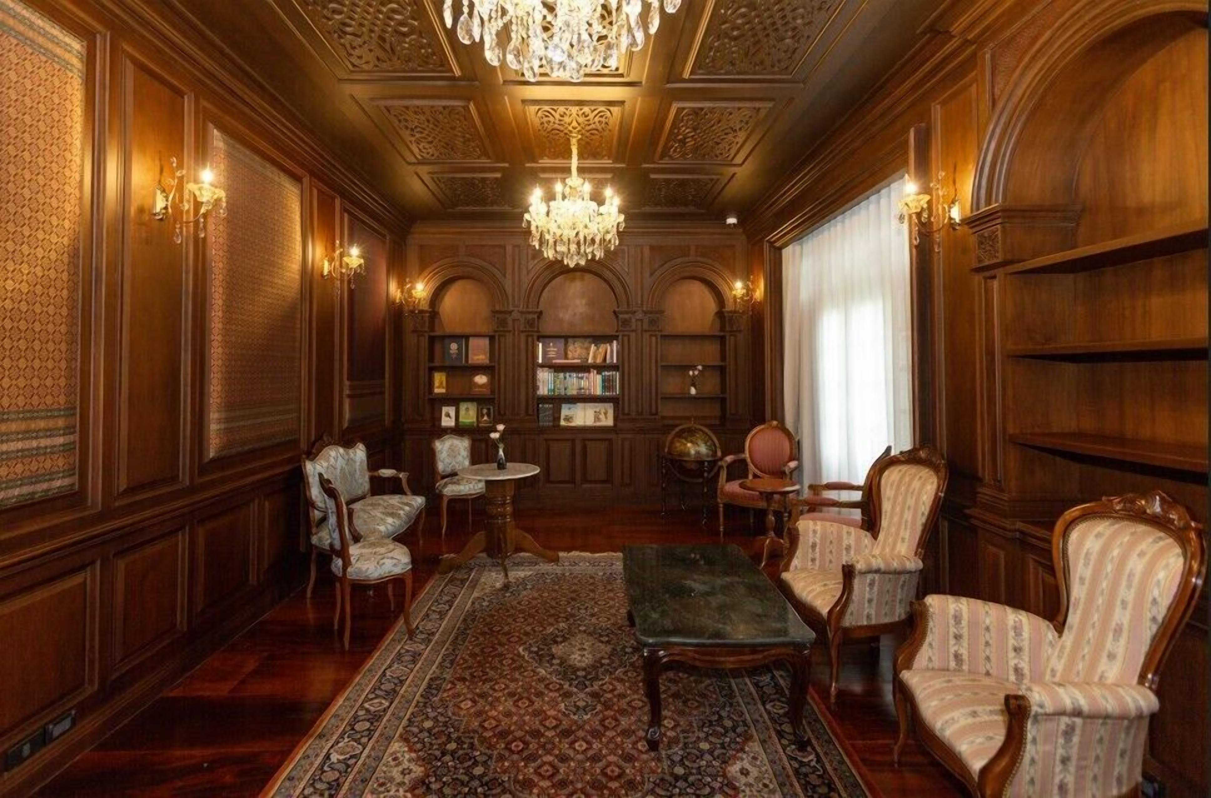 A room with ornate wood paneling, a chandelier, a bookshelf, and a patterned rug.