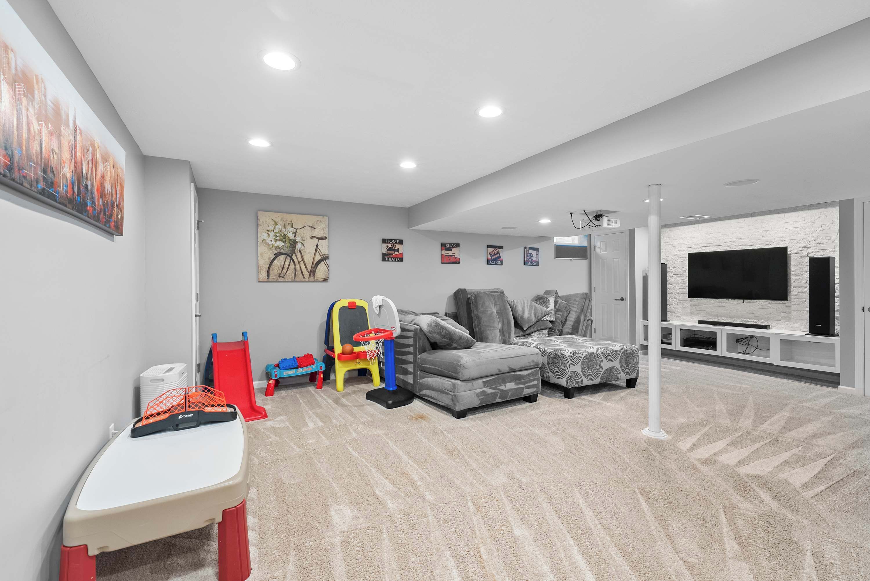 A modern living room with a grey couch, a small table, a red and yellow toy basketball hoop, and a large flat-screen TV mounted on a white brick wall.