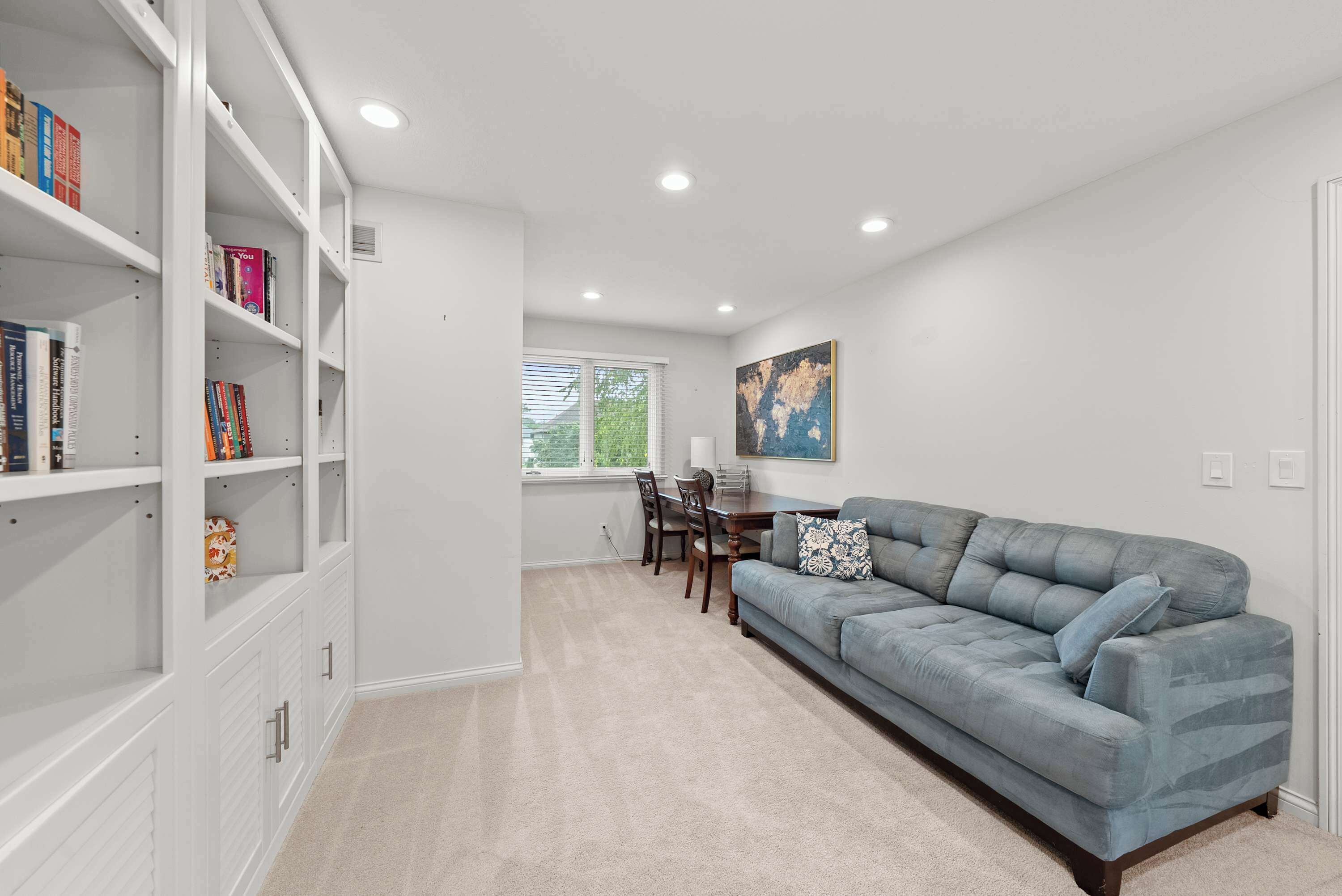 A spacious living room with a grey sofa, a bookshelf, a dining area with a table and chairs, and a window with blinds.