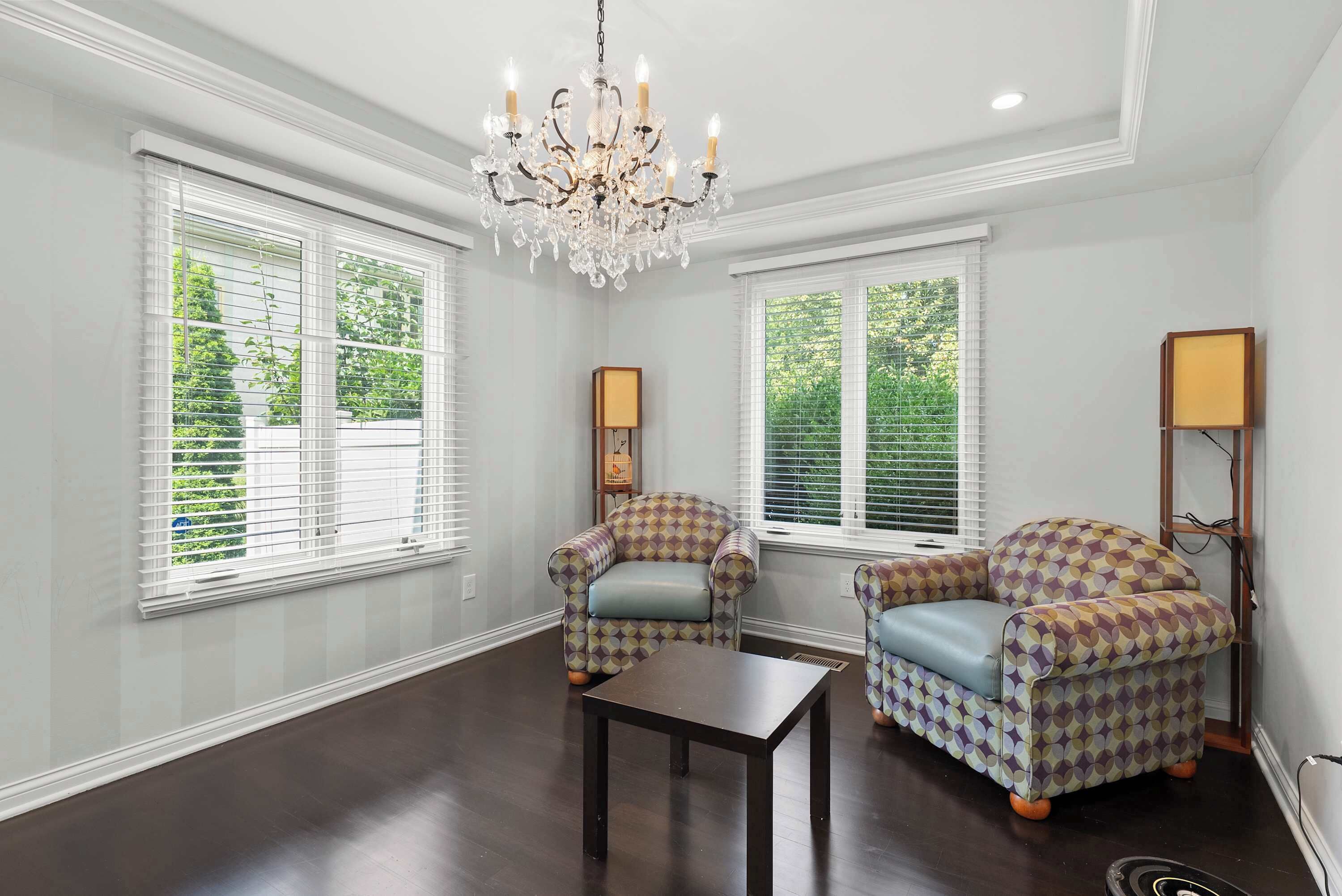A room with two patterned armchairs, a small table, and a chandelier. There are two windows with blinds, and a lamp is visible on the right side.